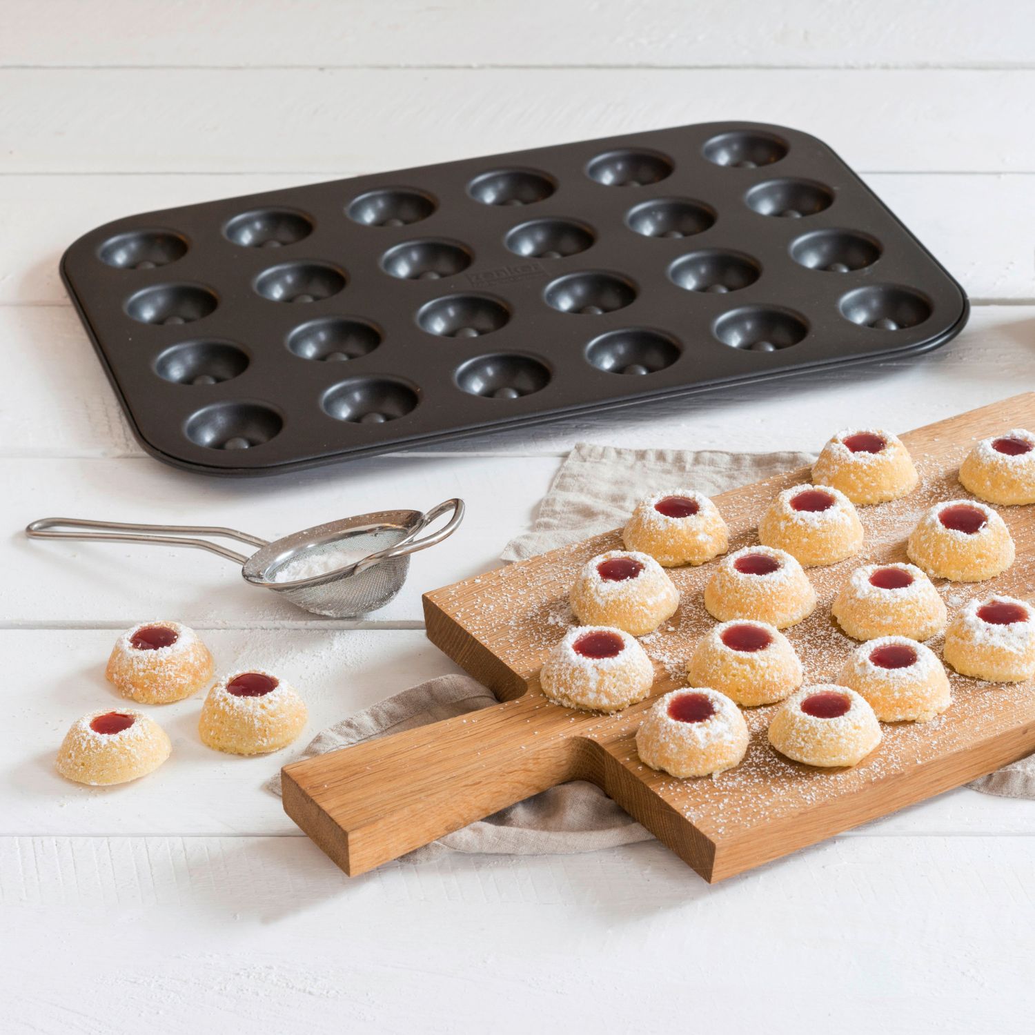 Husarenkrapfen auf einem Holzbrett vor Backblech für dieses Gebäck und neben Puderzuckersieb