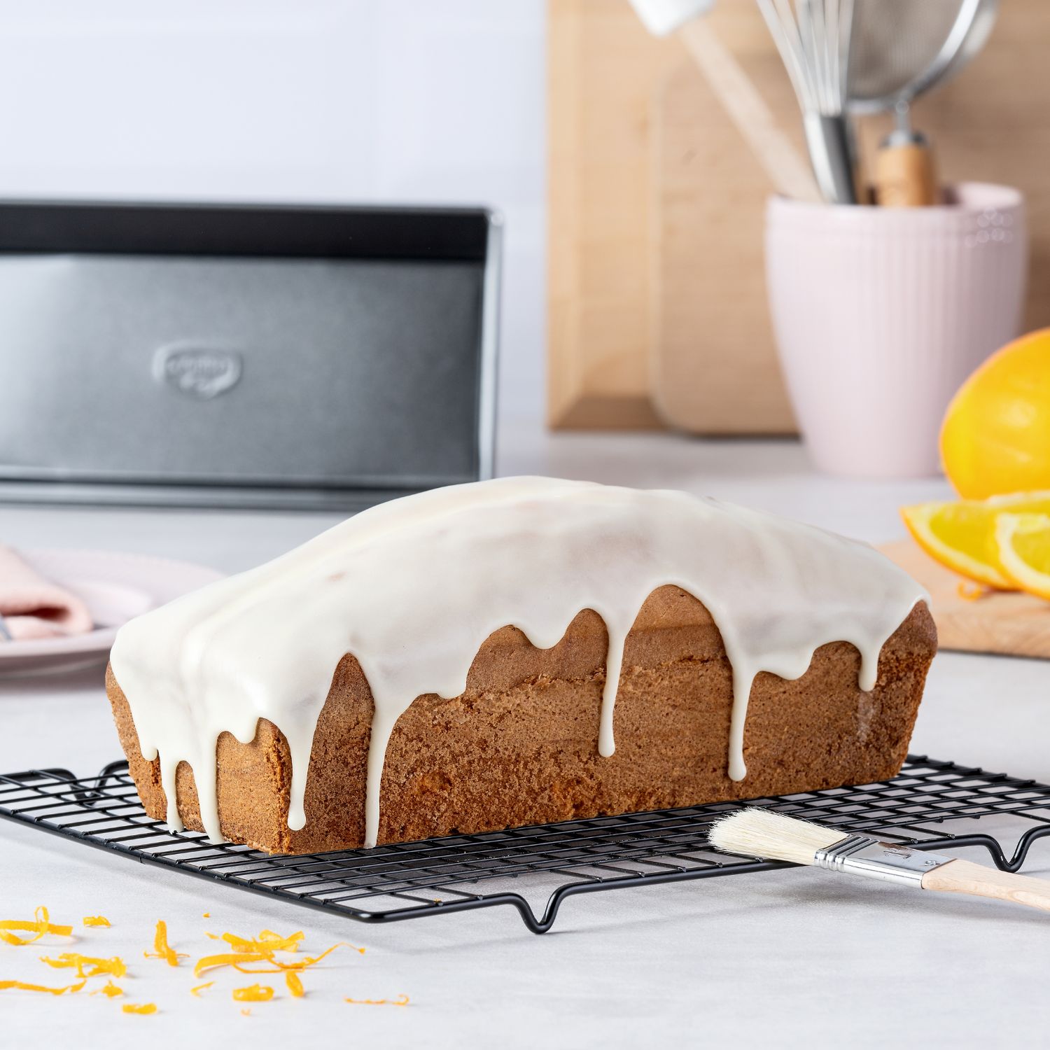 Ein Kasten-Kuchen mit weißer Glasur auf einem Backblech auf einer Küchen-Arbeitsplatte, Kasten-Backform liegt im Hintergrund