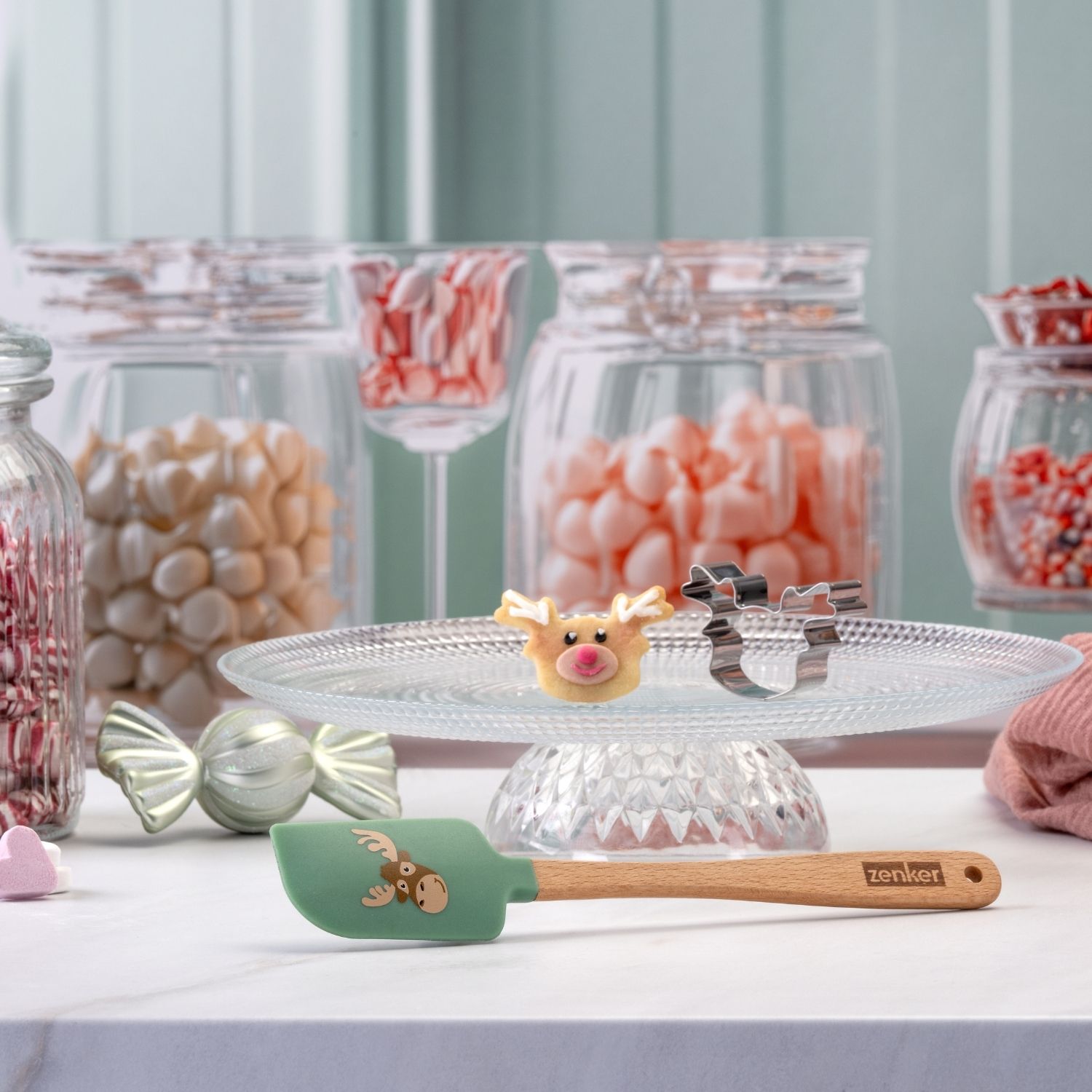 Mini-Teigschaber mit weihnachtlichem Motiv eines Elchkopfes plus Elchausstecher vor dekorativem Hintergrund mit Süßigkeiten