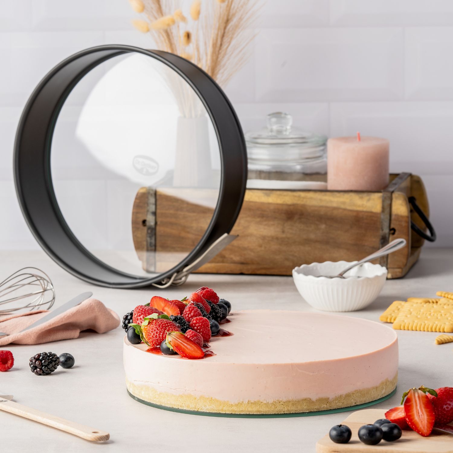 Küchen-Arbeitsplatte mit verschiedenen Backutensilien, einem Kuchen mit rosa Tortencreme und dekoriert mit Früchten, Springform steht im Hintergrund