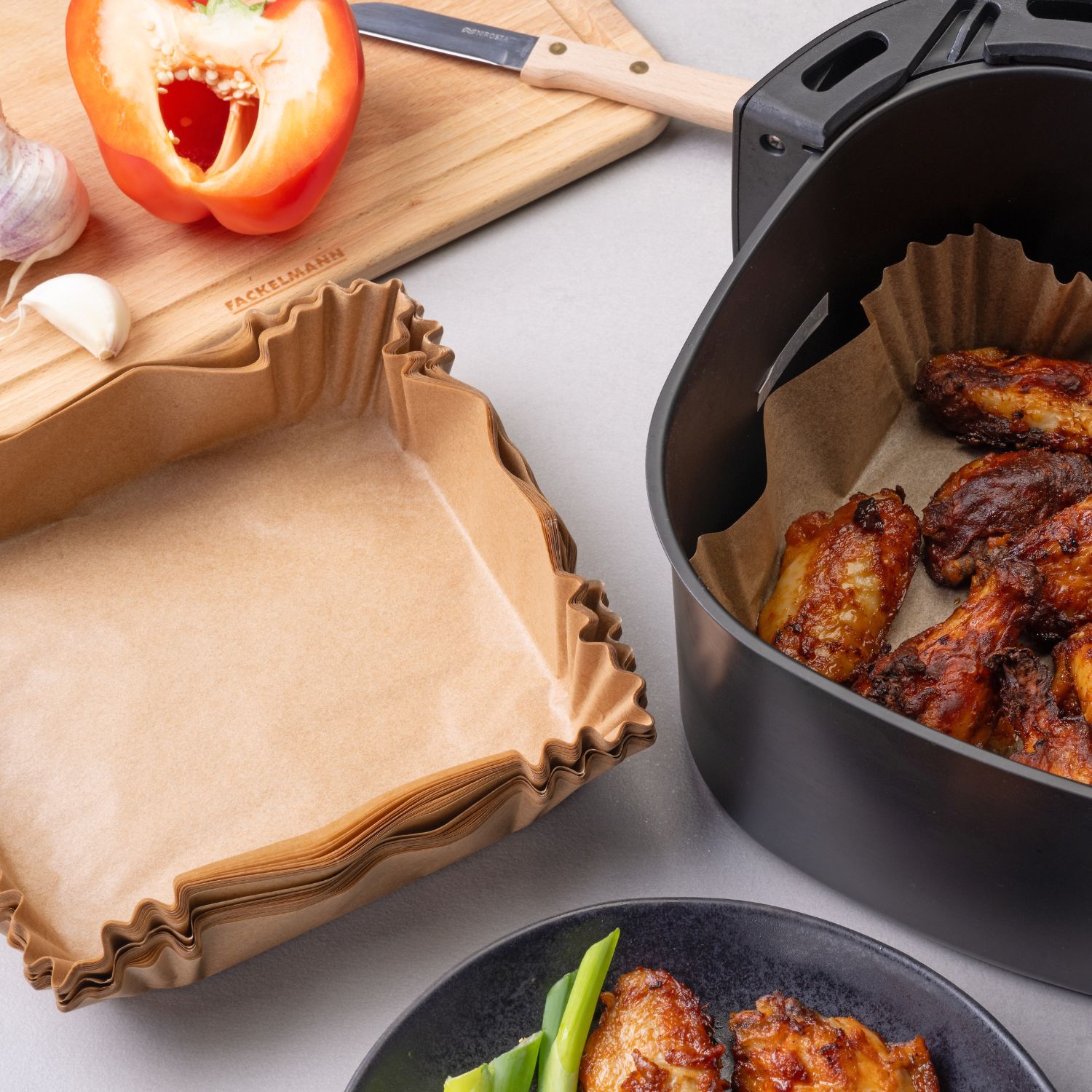 Eckiges hellbraunes Backpapier neben einer Heissluftfritteuse, die Chicken Wings enthält