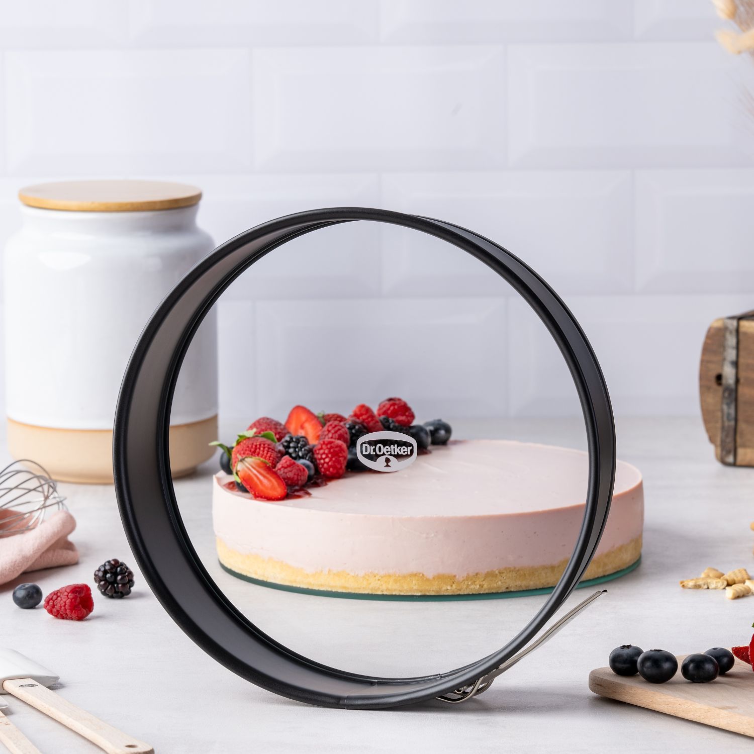 Springform auf einer Küchen-Arbeitsplatte mit verschiedenen Backutensilien und Torte im Hintergrund