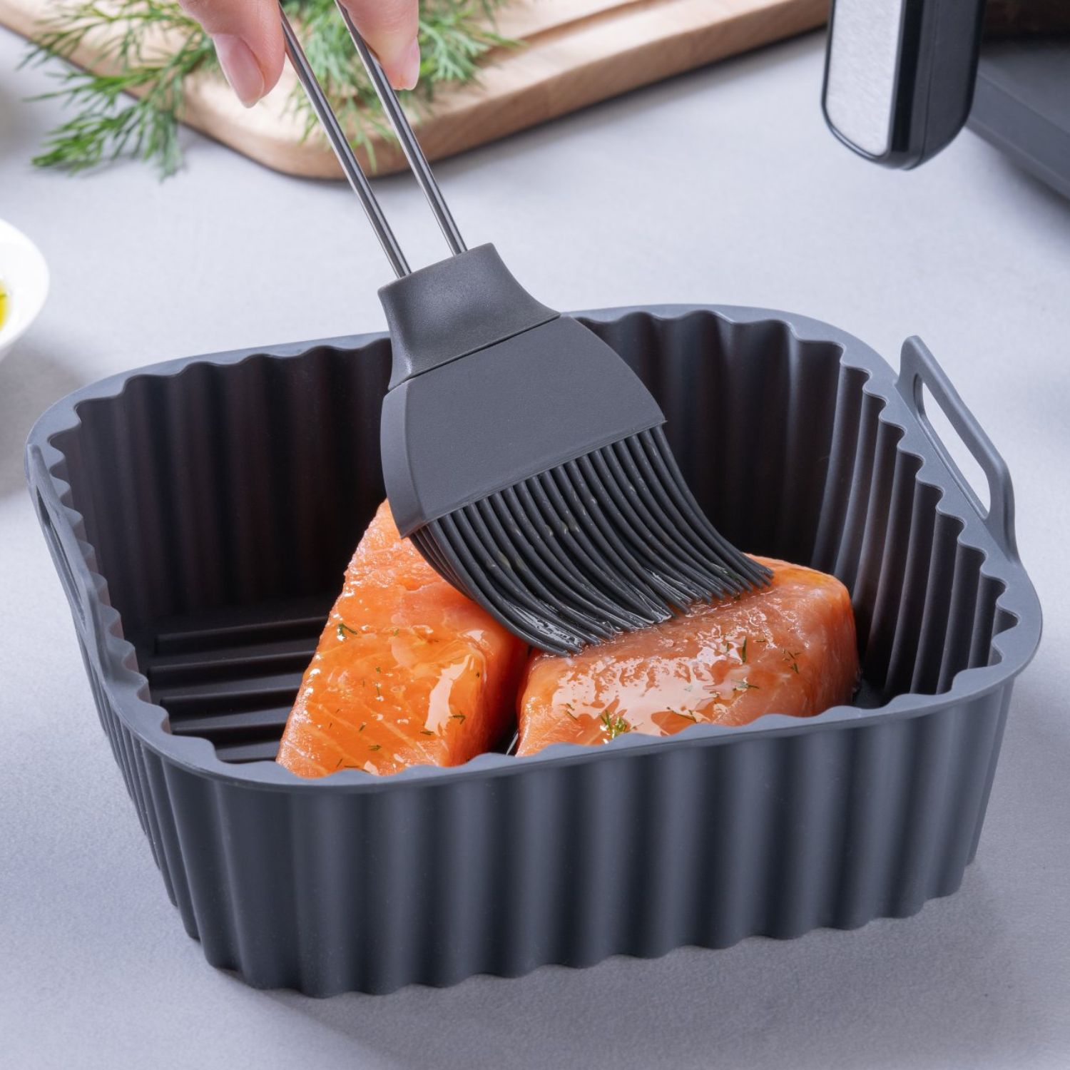 Eine eckige Silikonform für die Heißluftfritteuse mit rohem Lachs, auf den Öl mit einem Pinsel aufgetragen wird