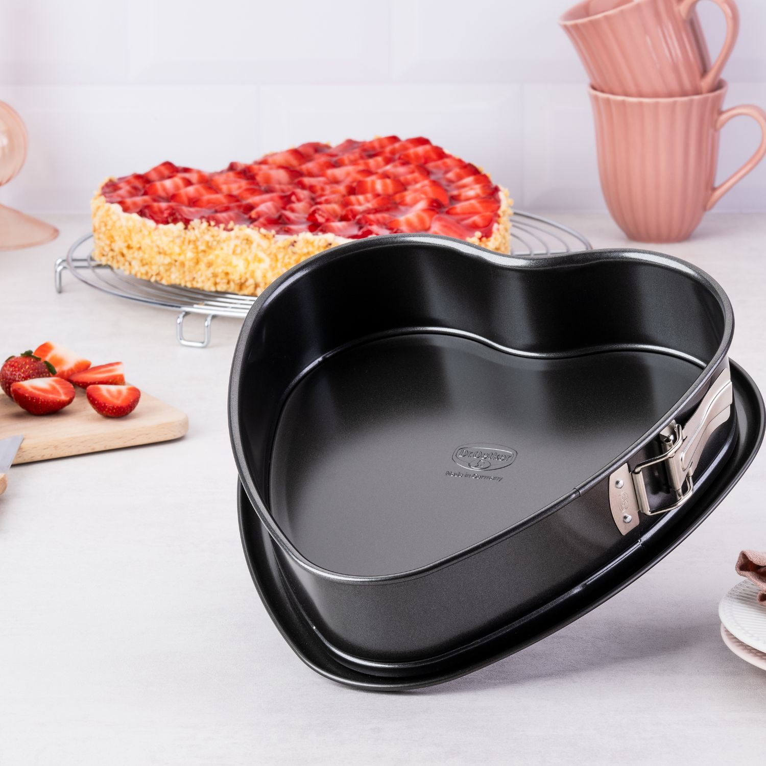 Eine schwarze Herz-Springform, im Hintergrund ein Herz-Erdbeerkuchen, Erdbeeren und pinke Tassen