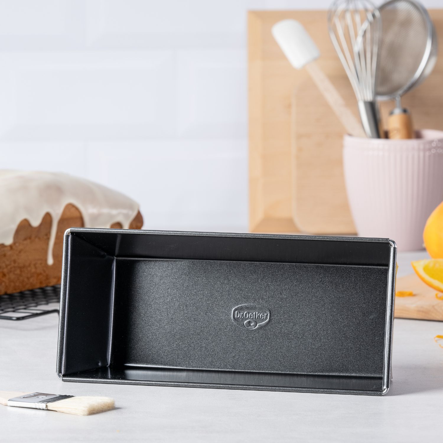 Eine schwarze Kasten-Backform mit Dr. Oetker Logo, liegend auf einer Küchen-Arbeitsplatte, mit Kuchen und Küchenutensilien im Hintergrund