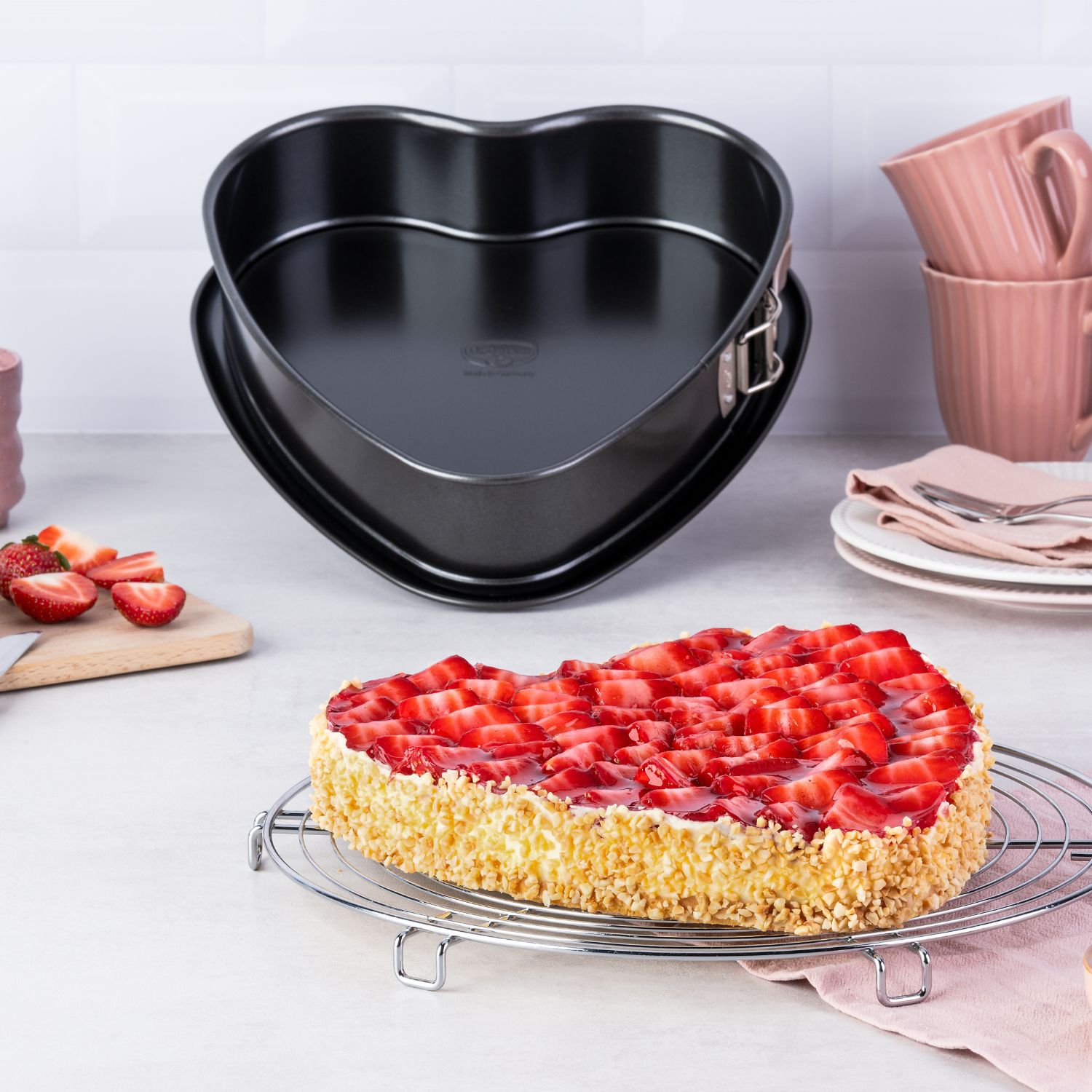 Ein Herz-Erdbeerkuchen im Vordergrund, dahinter die Herz-Springform, Teller, Erdbeeren und pinke Tassen