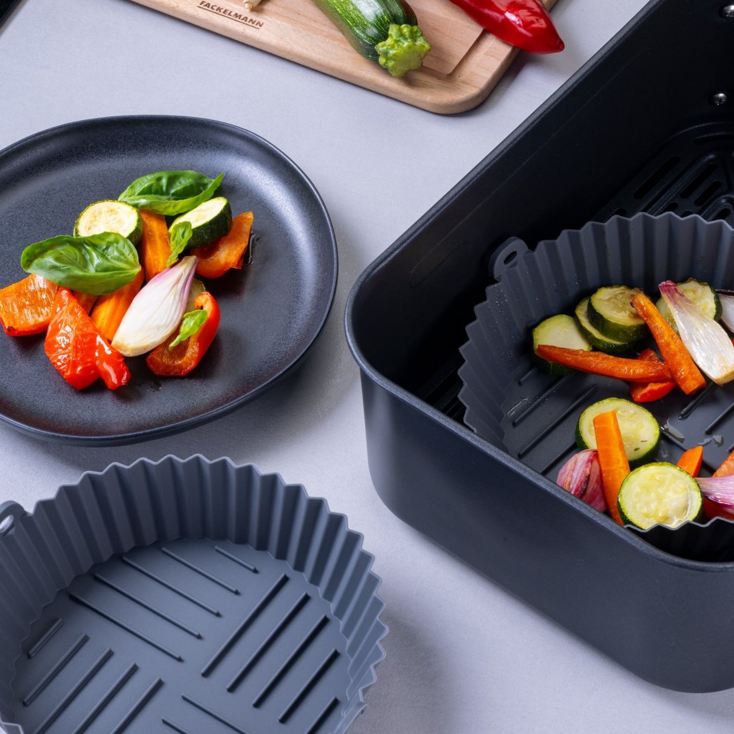 Zwei Silikonformen, eine befüllt mit Gemüse befindet sich in der Heißluftfritteuse, im Hintergrund steht ein Teller mit Gemüse