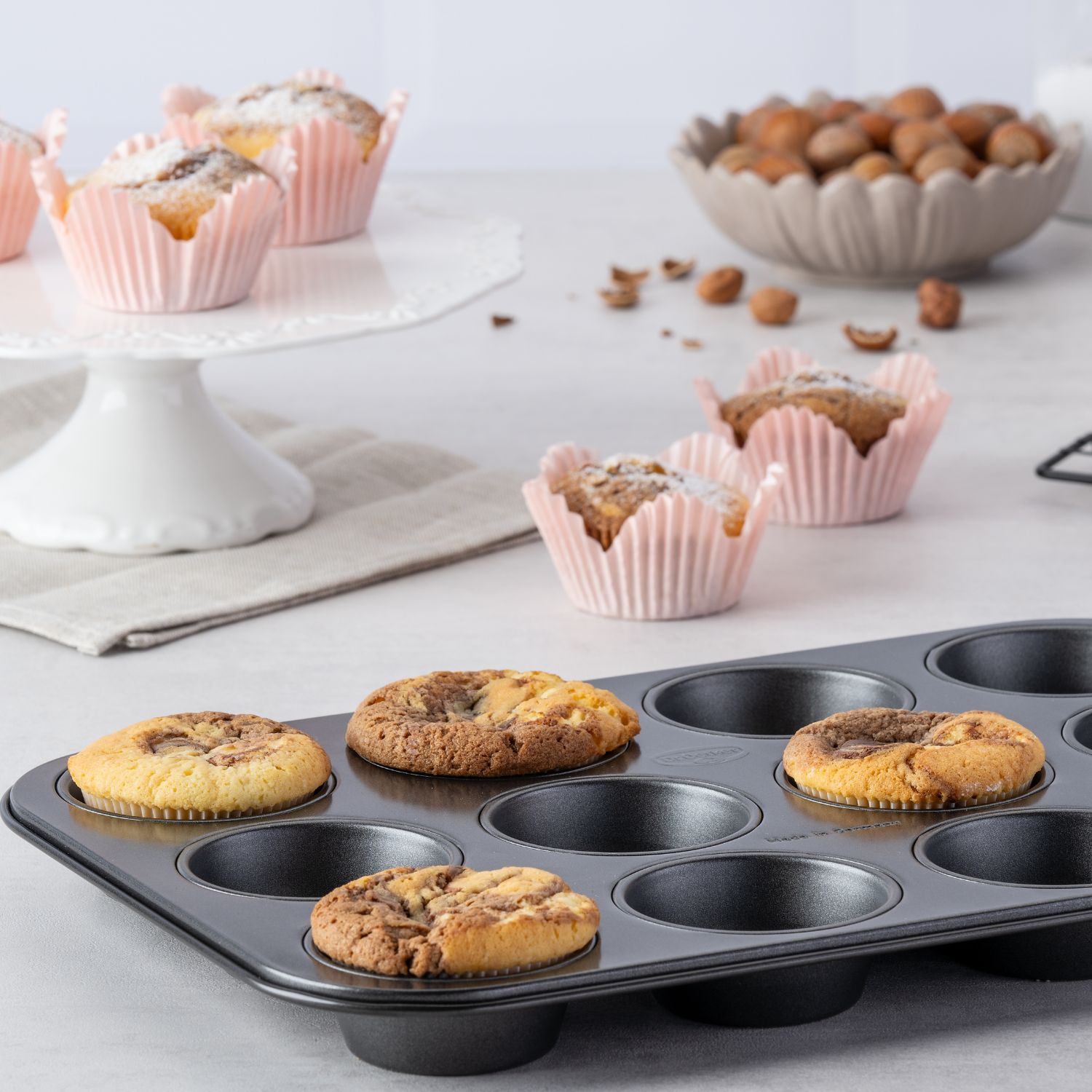 Ein schwarzes Muffinblech gefüllt mit vier Marmor-Muffins, auf einer Arbeitsplatte, Muffins in rosa Förmchen im Hintergrund