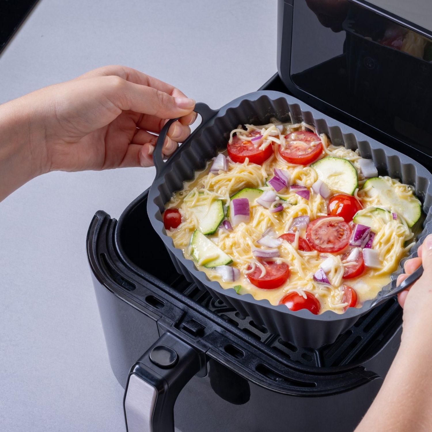 Eine eckige Silikonform die gerade in die Heißluftfritteuse geschoben wird, gefüllt mit einem Gemüse-Auflauf